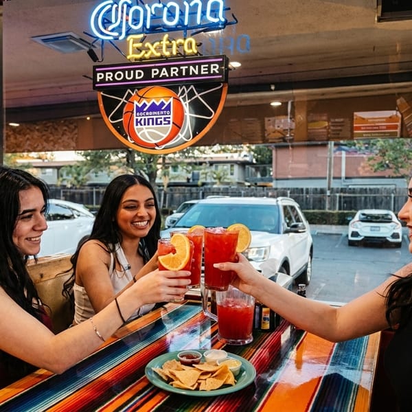 Three women toast red drinks with orange slices at a colorful restaurant table with chips and salsa.