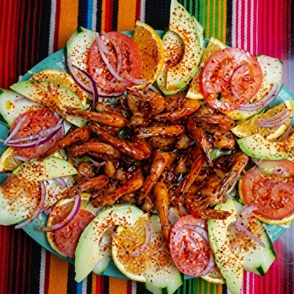 Shrimp dish with avocado, tomato, cucumber, and orange slices on a colorful striped tablecloth.