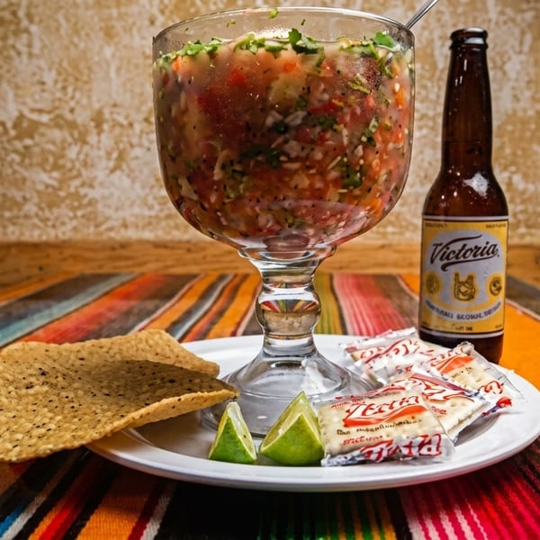 A large glass of ceviche with lime, crackers, and a Victoria beer on a colorful striped tablecloth.
