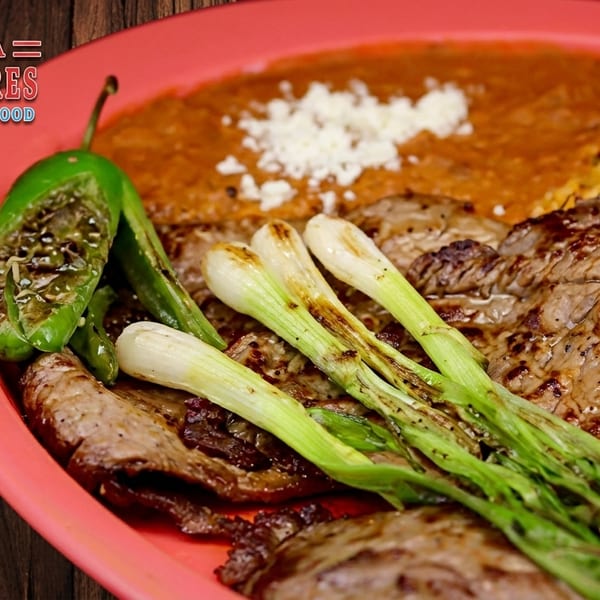 Grilled steak, charred green onions, and jalapeños with rice and beans on a red plate.